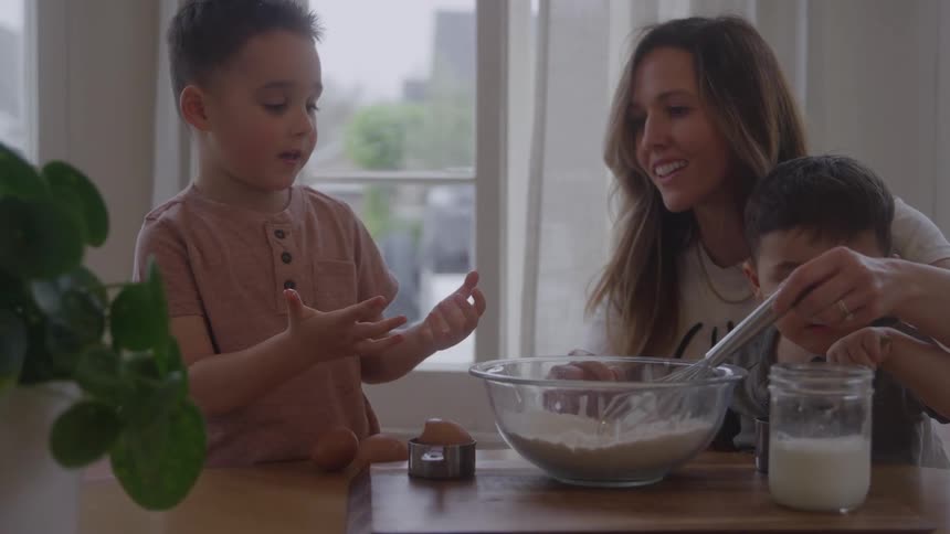 A woman and two young boys stand around a kitchen counter one boy helps the woman whisk ingredients in a glass bowl the other boy stands and looks at his hands a large window provides soft background lighting