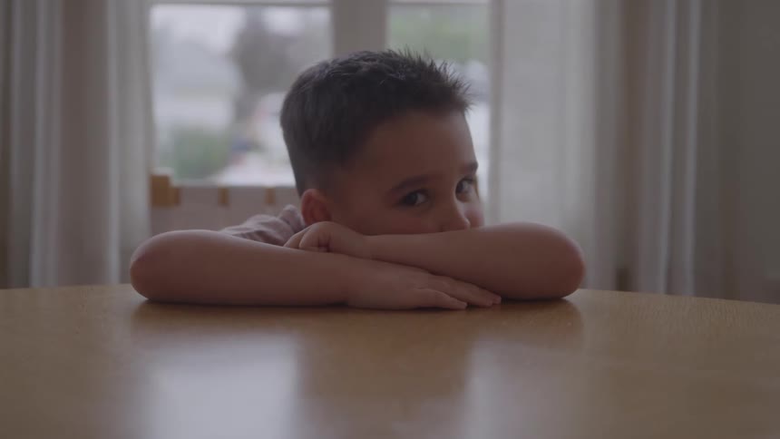 A young boy rests his head on crossed arms on a wooden table looking up with a neutral expression toward the camera with a window and soft light in the background