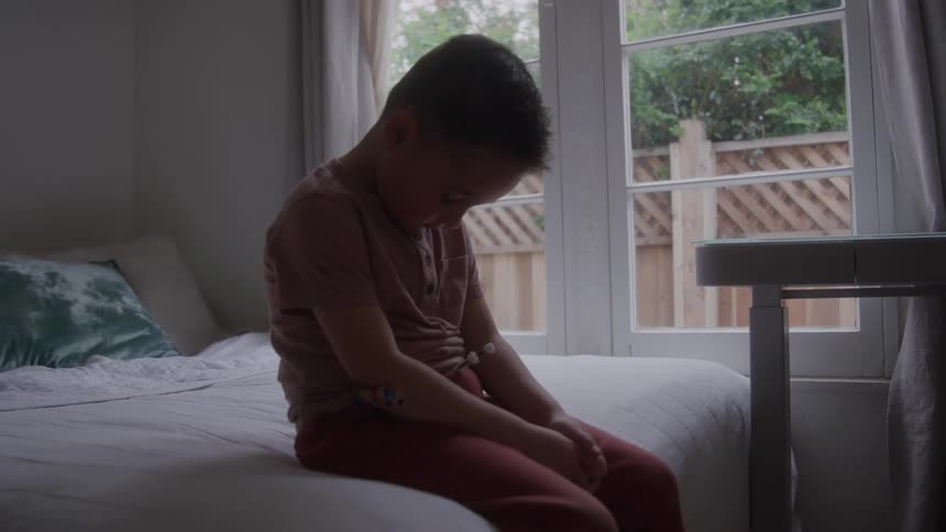 The video is an interior shot of a young boy sitting on a bed in a room with a large window he is facing the window with his head bowed and arms resting on his knees conveying a sense of quiet contemplation or sadness