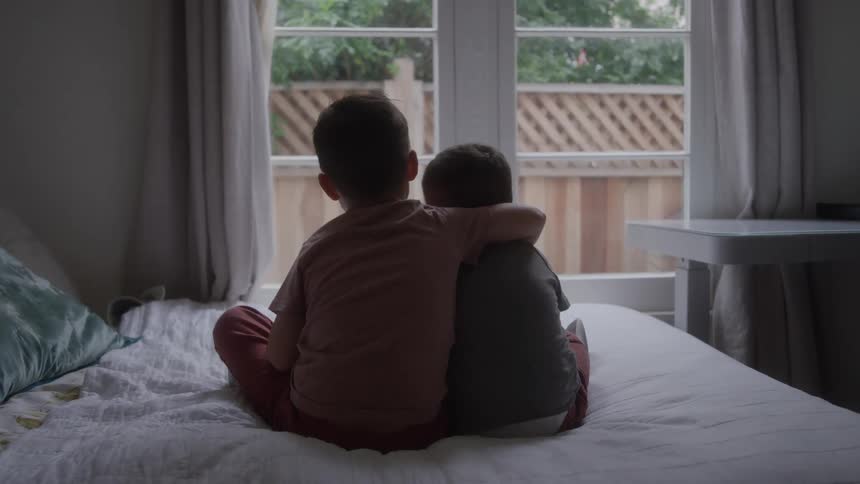 Boy puts an arm around his younger brother while sitting on the bed with him