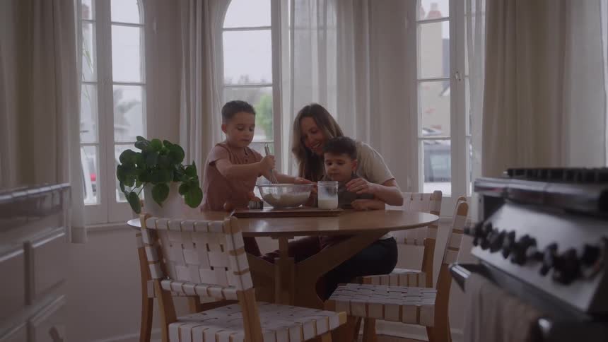 Mother smiles while watching one of her two sons stirring a mixture as they sit at the breakfast table