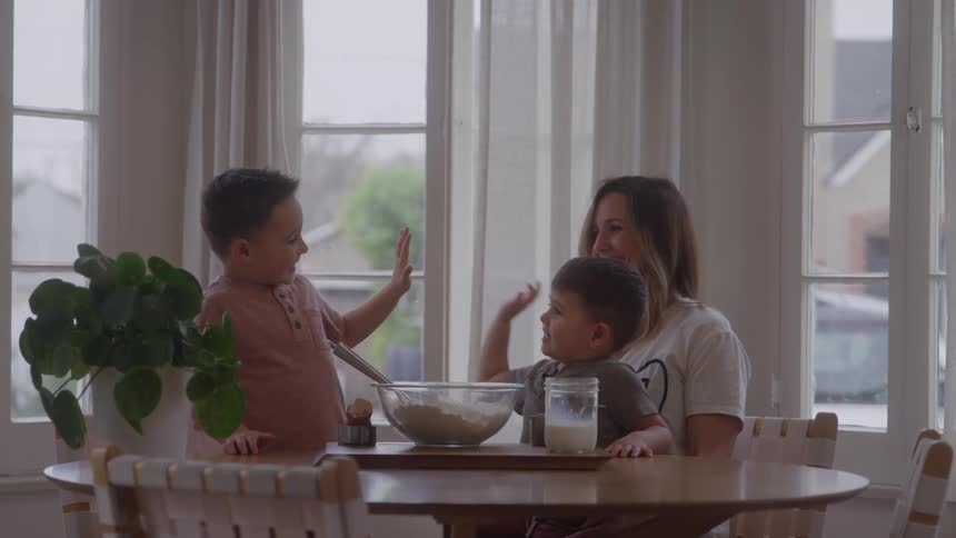 A happy mother sits at a dining table with her two young sons who playfully high five each other while preparing to bake