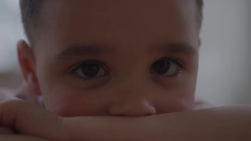A closeup of a young boys face the boy rests his chin on his forearm only the upper portion of his face is clearly visible the eyes look directly at the camera with a searching gaze the lighting is dim and ambient