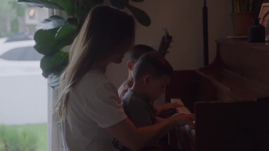 A woman and two boys sit at a dark upright piano the boys are playing the keys while the woman assists them a large window behind them illuminates the subjects from the side a large potted plant stands in the foreground