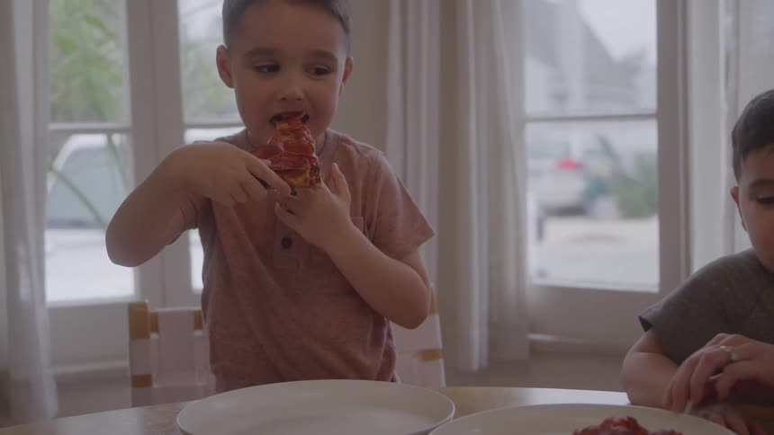 Two young boys eat pizza at a table in a bright home dining area