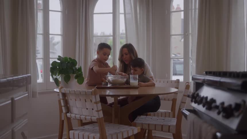A family gathered around a table baking a cake the mother holding a small child in her arms and another child mixing the cake batter with a whisk
