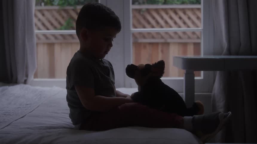 A small boy sits crosslegged on a bed the boy holds a stuffed toy in his lap the subject is silhouetted against a large window light filters in from the outside the scene is set inside a bedroom