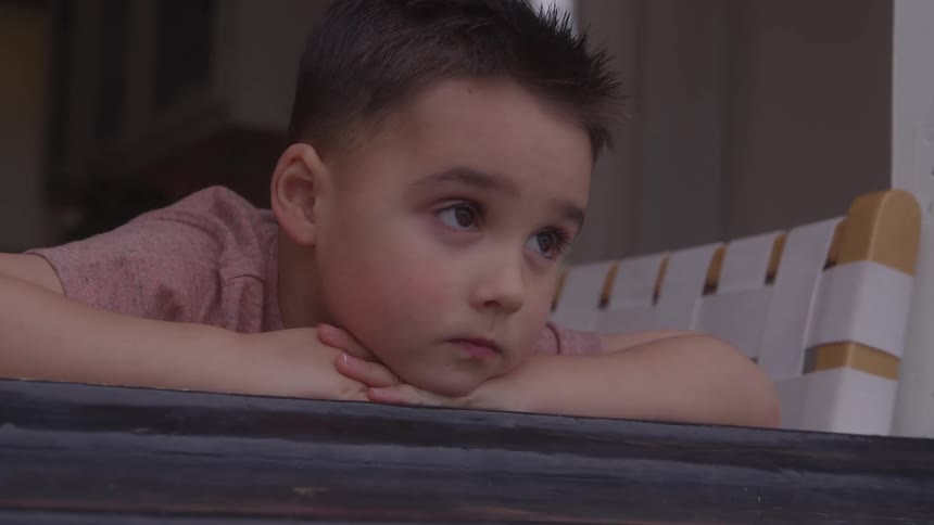 A young boy rests his chin on his arms the boy looks out a window he has a contemplative expression a white and yellow bench sits in the background