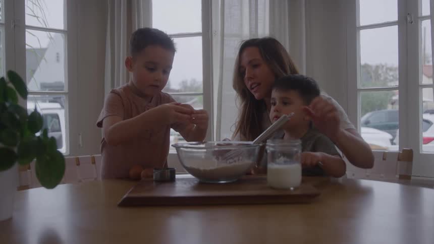The video shows a woman and two young boys standing at a table next to a large window suggesting a bright kitchen setting the woman is actively helping the children whisk ingredients in a bowl indicating they are baking or cooking together