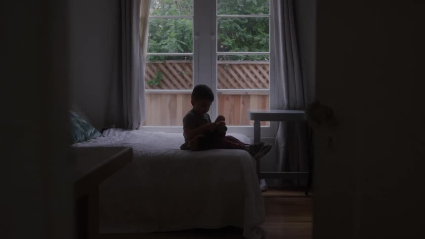 A silhouetted small child is sitting on a white bed holding a stuffed animal or doll while looking toward a bright window with greenery visible outside framed by a dark doorway