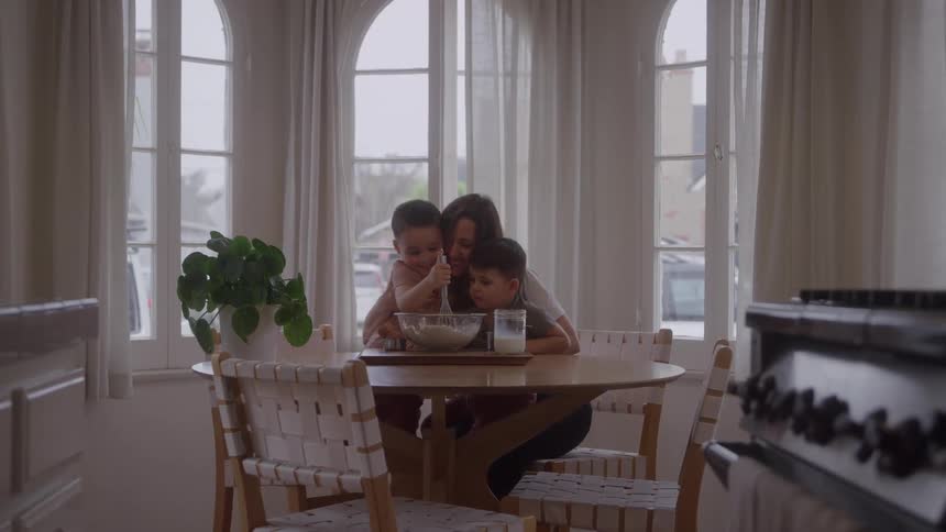 A woman sits at a round kitchen table with two young boys as they stir a mixing bowl together by tall windows