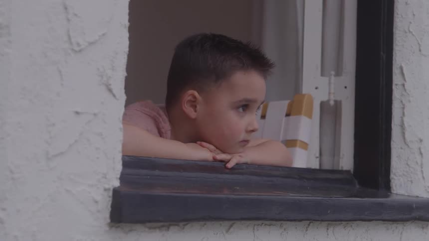 A close view from outside shows a young boy with dark hair resting his chin on his hands looking out pensively from a small dark framed window set in a rough white wall