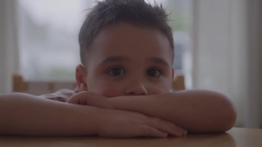 A young boy with wide expressive eyes rests his head on his arms at a table