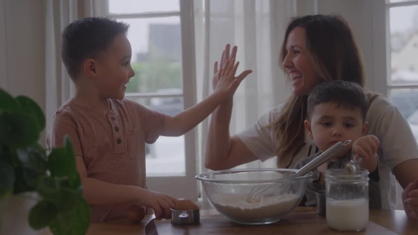 A woman and two young boys are gathered around a wooden counter the woman and the taller boy share a highfive and smile the smaller boy stirs a liquid in a jar a glass bowl of flour is placed between them a large window lights the scene