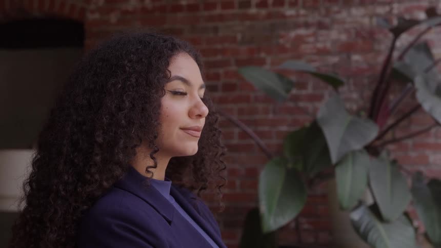 A woman with curly hair in a navy blazer looks confidently to the side against a brick wall and leafy plant