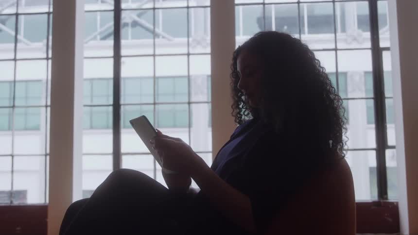 The video is an interior shot of a woman silhouetted against a large gridpaned window as she sits and looks at a tablet the contrast between the bright window and her dark silhouette emphasizes the loftstyle setting and her concentration on the device