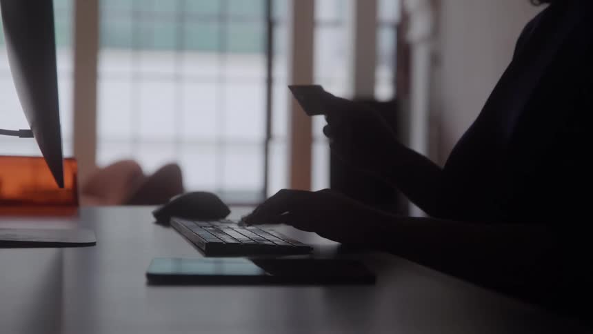 Silhouetted against a bright window a person makes an online purchase by holding a credit card and typing on a computer keyboard