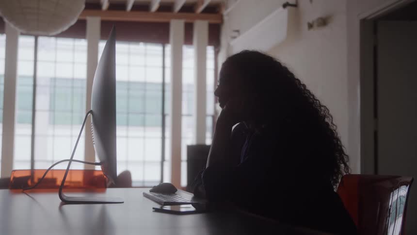 The video shows a woman seated in a chair working on a computer in a brightly lit office or home office with a large window she is seen in silhouette which highlights the light pouring in from the windows behind her