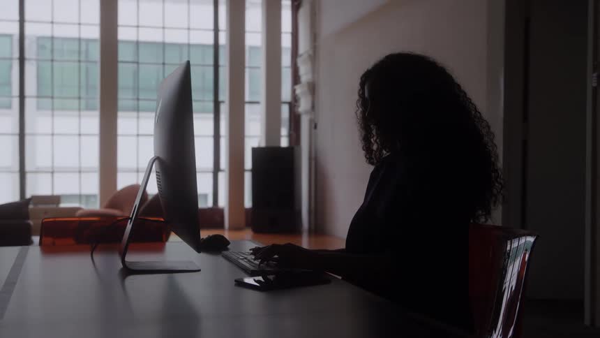 A woman is silhouetted at a desk typing on a computer in a bright loft with large windows