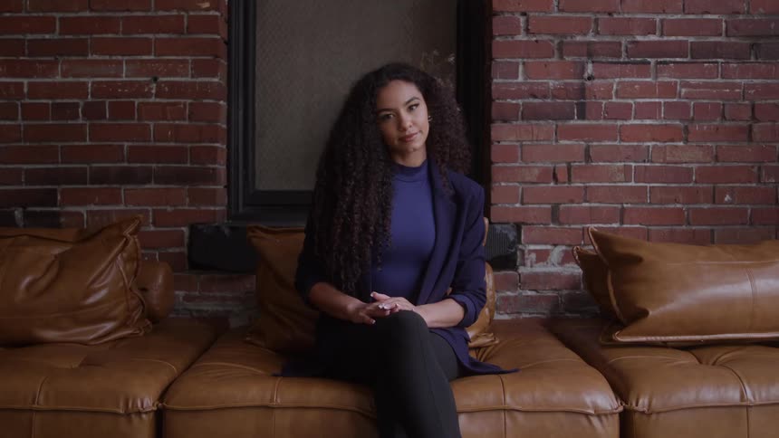 A woman sits centered on a brown leather couch facing the camera the woman wears a dark blue blazer and pants the background consists of a red brick wall with a dark rectangular window opening