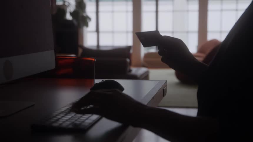 A closeup of a persons hands shows one hand typing on a keyboard the other hand holds a small plastic card the person and desk are silhouetted against a bright background window