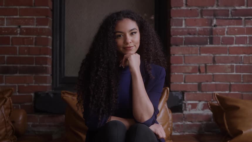 The video shows a medium shot of a young woman with long curly hair sitting indoors against a dark brick wall she is looking off to the side with a gentle thoughtful expression before turning to face the camera with a slight smile