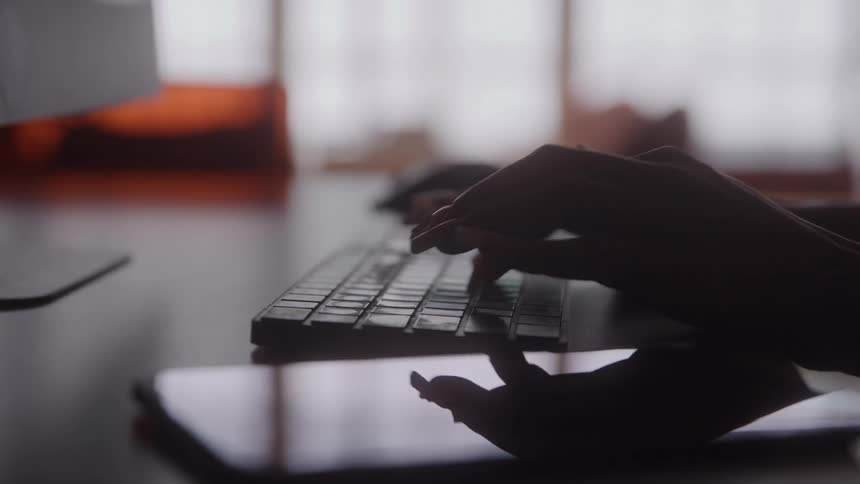 A closeup of hands typing on a keyboard at a desk in a dimly lit workspace