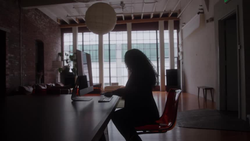 Person seated on transparent chair working on computer in office with large windows and industrial decor