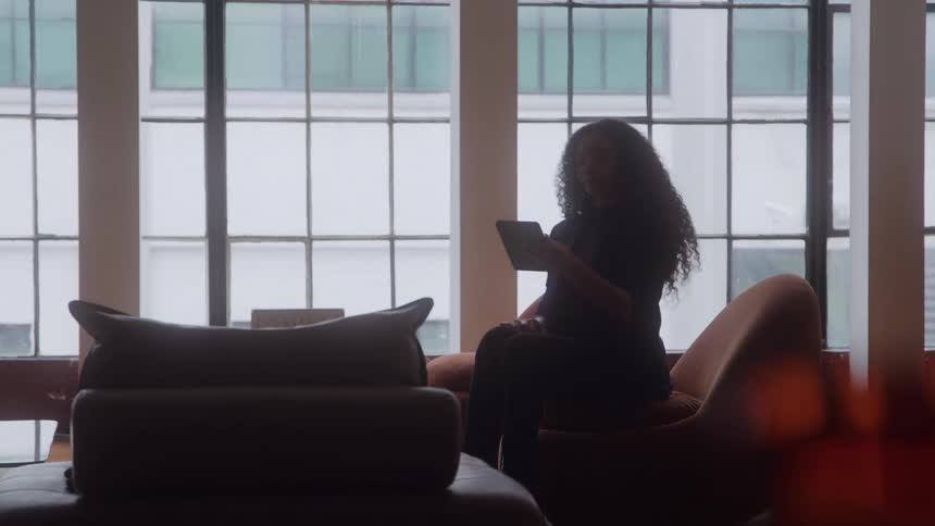 Woman walks into a room holding a tablet computer and sits down in a chair