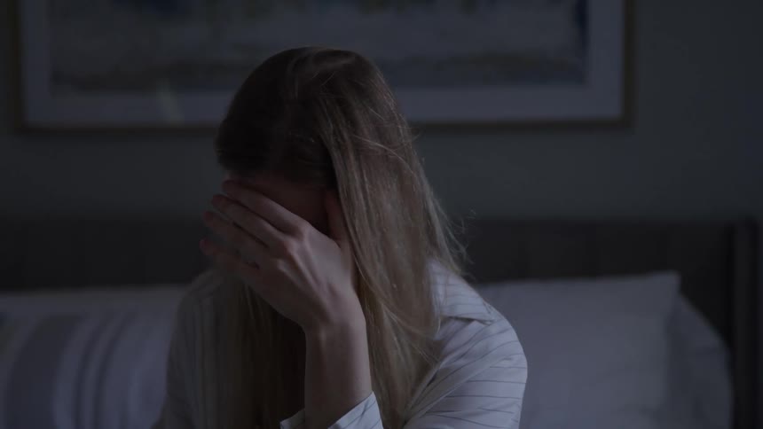 Blonde woman sitting on her bed looks toward the sunlight before covering her eyes with her hand