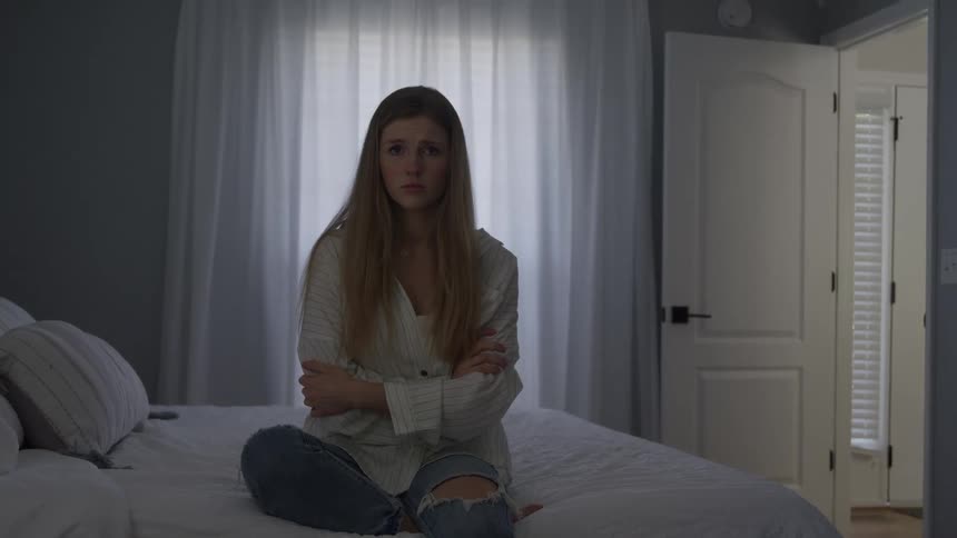 A sad looking woman with long brown hair wearing a white shirt and ripped jeans sits on a white bed with her arms crossed in a darkened bedroom