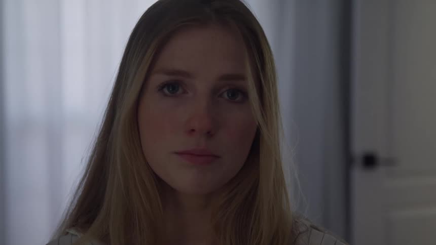 Blondehaired woman looking straight ahead in indoor setting with curtains and door in the background