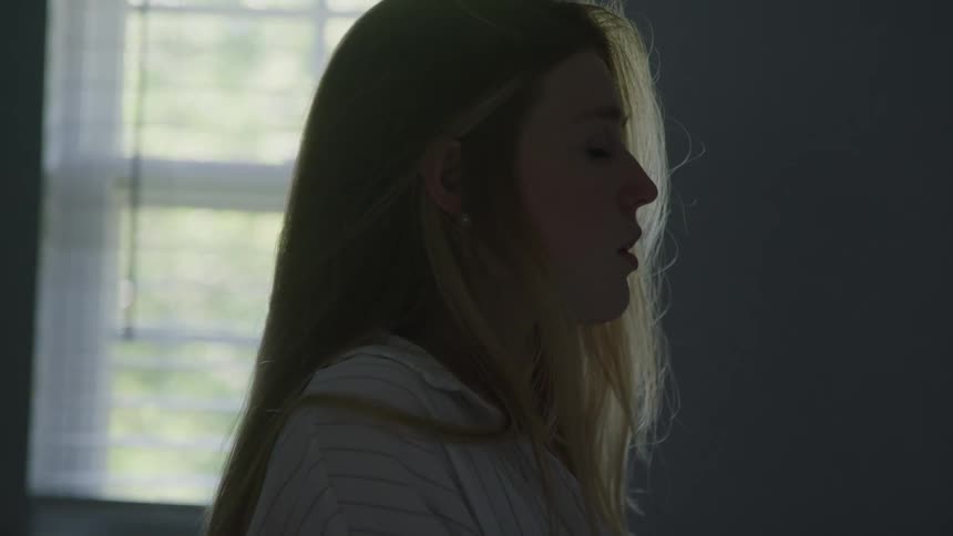 A woman stands in a dim room lowers her head then slowly raises her head to look forward light comes from a window behind her creating a silhouette effect