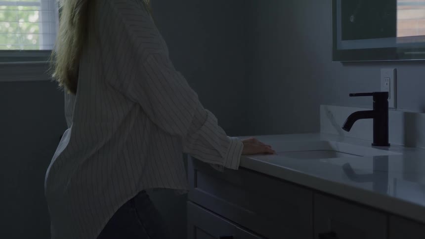 Blonde woman standing with her hand on the sink counter drops her head forward