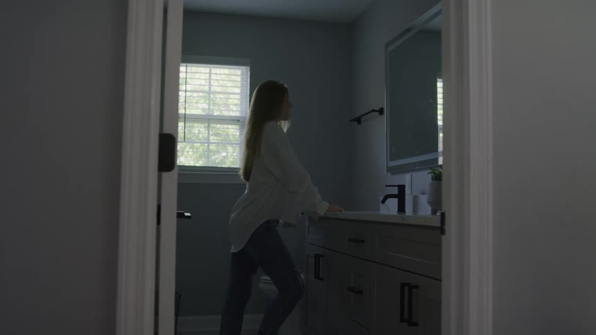 Woman standing at sink in modern bathroom with gray wall and white shuttered window