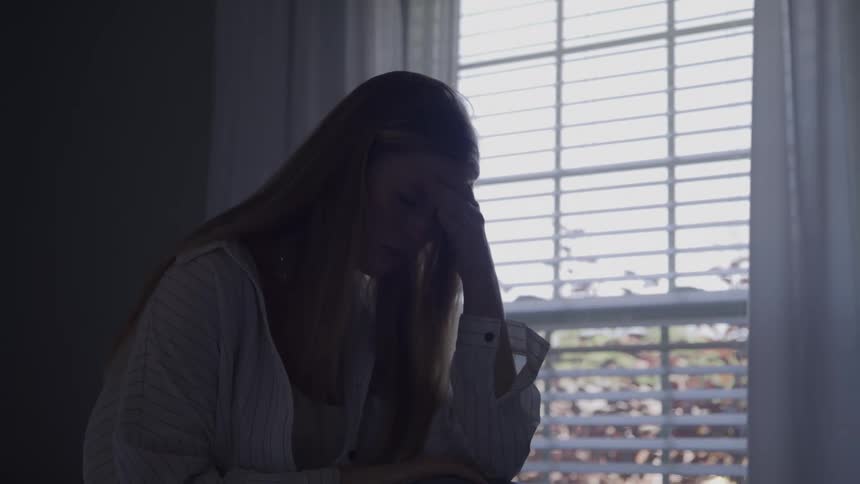 A depressed woman sits by a window in a dimly lit room
