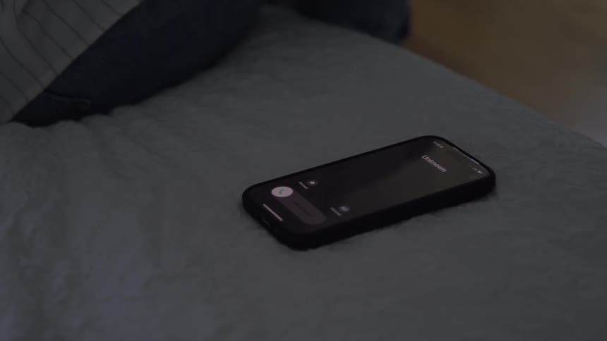 A hand reaches for a smartphone on a bed in a dimly lit bedroom