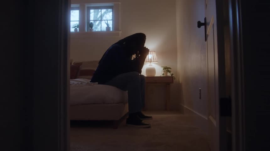 Man sitting on edge of bed with hands clasped near face in room lit by table lamp