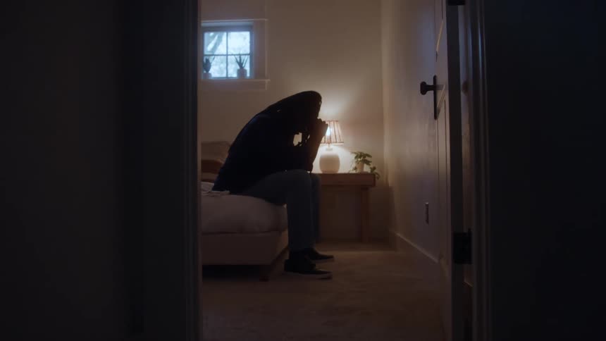 A silhouetted depressed person sits on a bed in a dimly lit bedroom