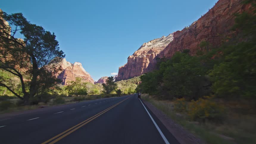 A highway road winds through canyon cliffs and trees under a clear blue sky