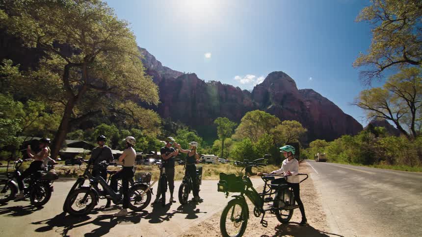 A group of cyclists all wearing helmets are parked along the roadside engaged in conversation under the bright midday sun
