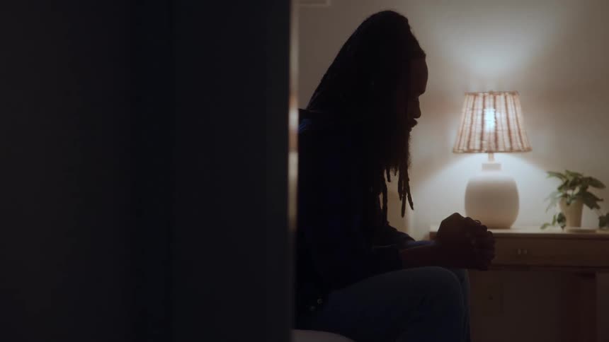 A man sitting on a bed in a bedroom with his hands together praying