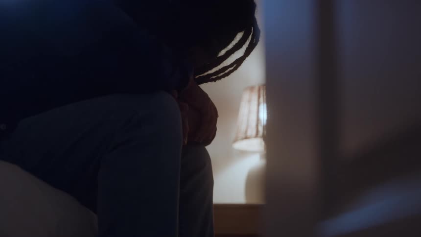 A silhouette of a person wearing dark clothing and light pants is slumped over sitting alone in a dim room with a glowing lamp visible in the background