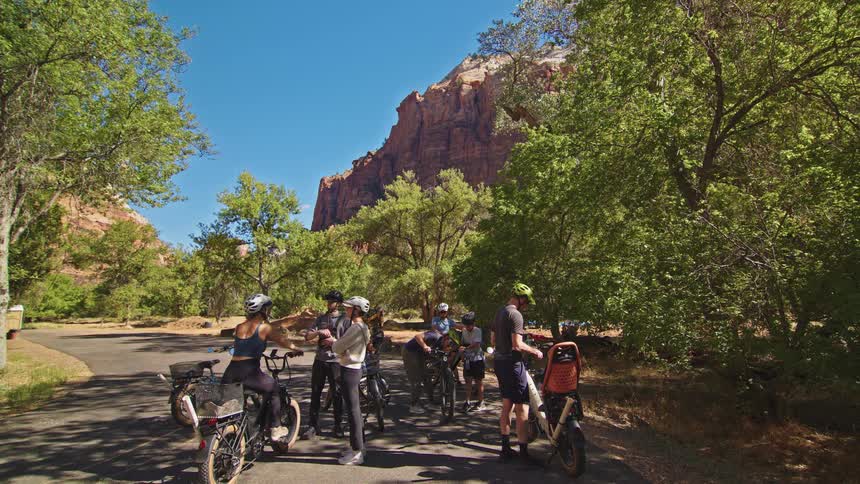 Group of cyclists gathered on paved trail surrounded by trees and red rock formations