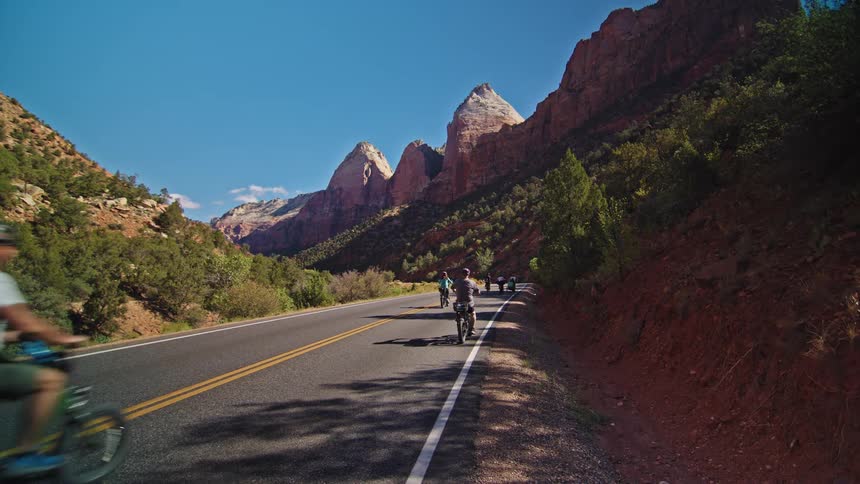 Cyclists riding along road flanked by green vegetation and rocky mountains under blue sky