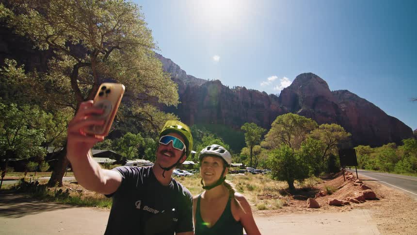 A group of cyclists are parked along the roadside on a sunny day all wearing helmets while a young man and woman from the group take a selfie together