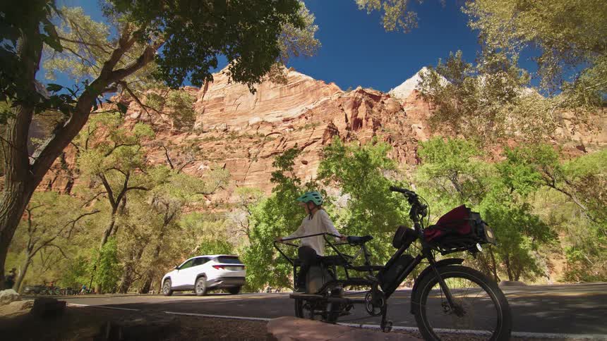 Cyclist takes a break by the roadside surrounded by trees and rocky mountains