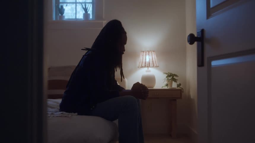 A dimly lit interior view through a doorway shows a silhouetted woman sitting alone on a bed looking down sadly with her hands clasped next to a glowing bedside lamp