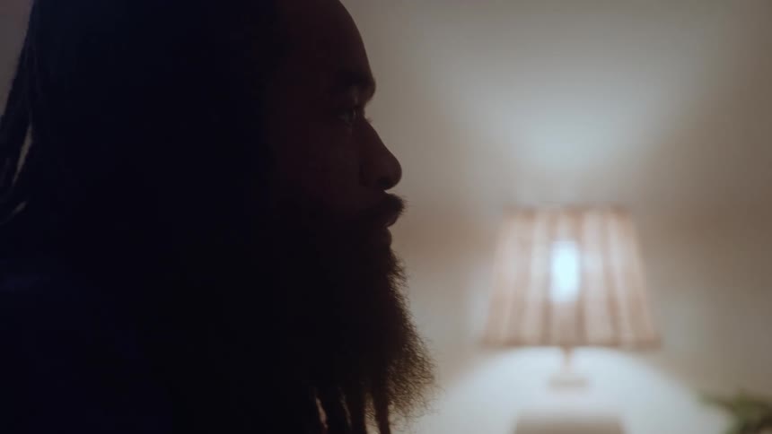 A silhouette close view shows a man with long dark dreadlocks and a beard standing in a dimly lit room with a soft glowing table lamp and a potted plant blurred in the background