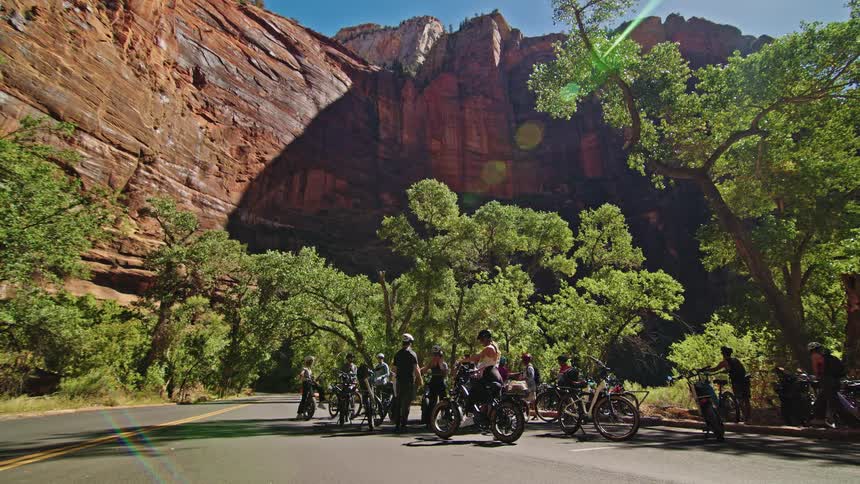 A group of bikers gather along the side of a freeway surrounded by mountains under a clear sunny sky